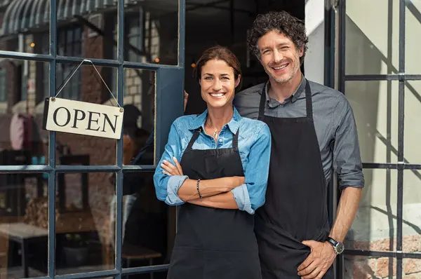 Two cheerful small business owners smiling and looking at camera while standing at entrance door. Happy mature man and mid woman at entrance of newly opened restaurant with open sign board. Smiling couple welcoming customers to small business shop.